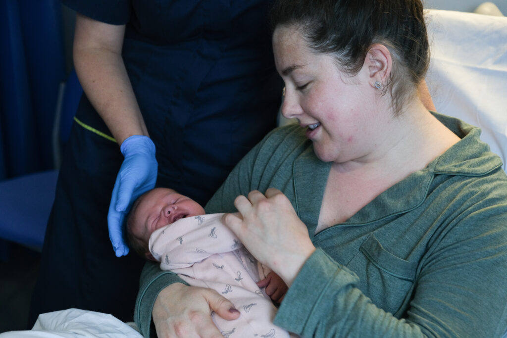 A photo of a woman gazing down at her newborn baby. The baby is dressed in a pink outfit. A midwife's gloved hand is in shot cradling the baby's head. 