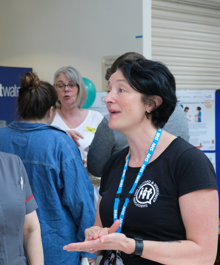 A photo of a woman in a black tshirt with the Maternity and Neonatal Voices logo on it. She is wearing an NHS lanyard and has short black hair. She is in the middle of talking and groups of people are in the background. 