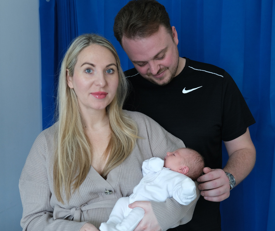 A photo of a white woman with blonde hair dressed in a cardigan holding a newborn baby in a white outfit. A man stands next to them in a black tshirt and gazes down at the baby smiling.